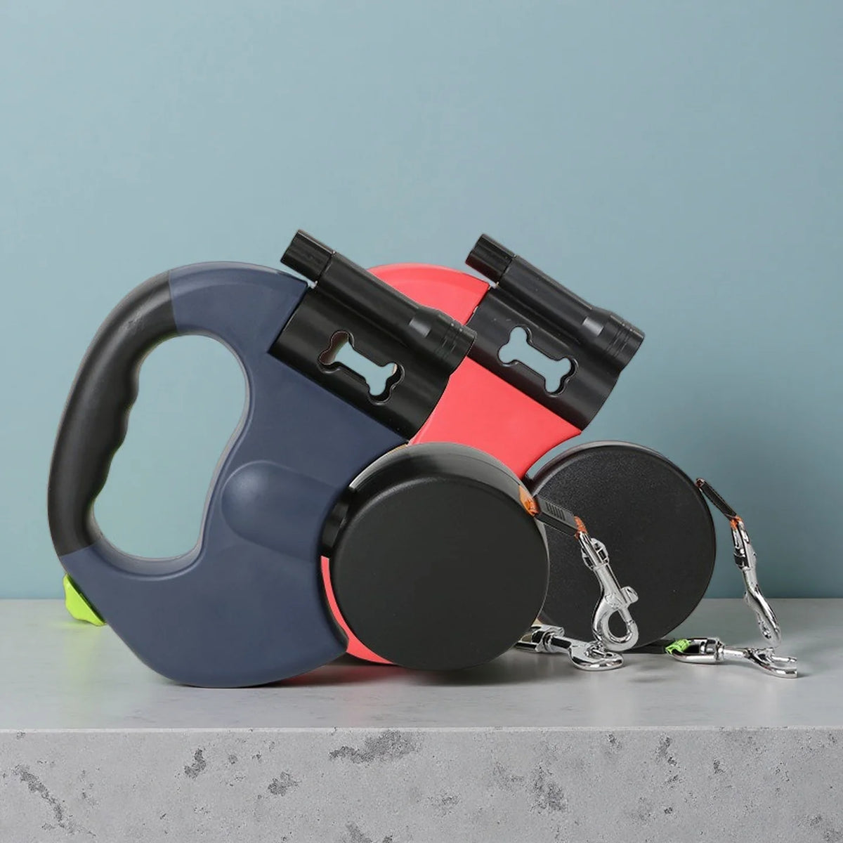 A black and gray retractable dual dog leash with a red handle and LED light is displayed on a gray surface.