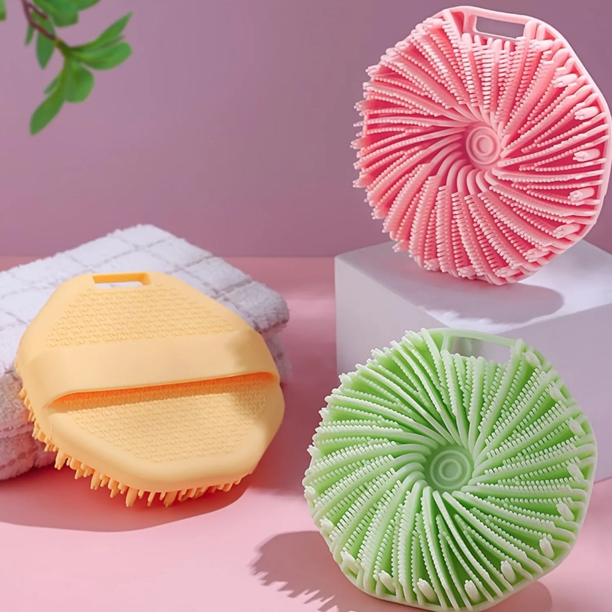 A set of soft silicone exfoliating brushes in pink and green are displayed on a pink surface.