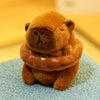 Flauschiger brauner Capybara-Plüschtier mit Hoodie und kleinen Flügeln, sitzt auf einer hellblauen Oberfläche.