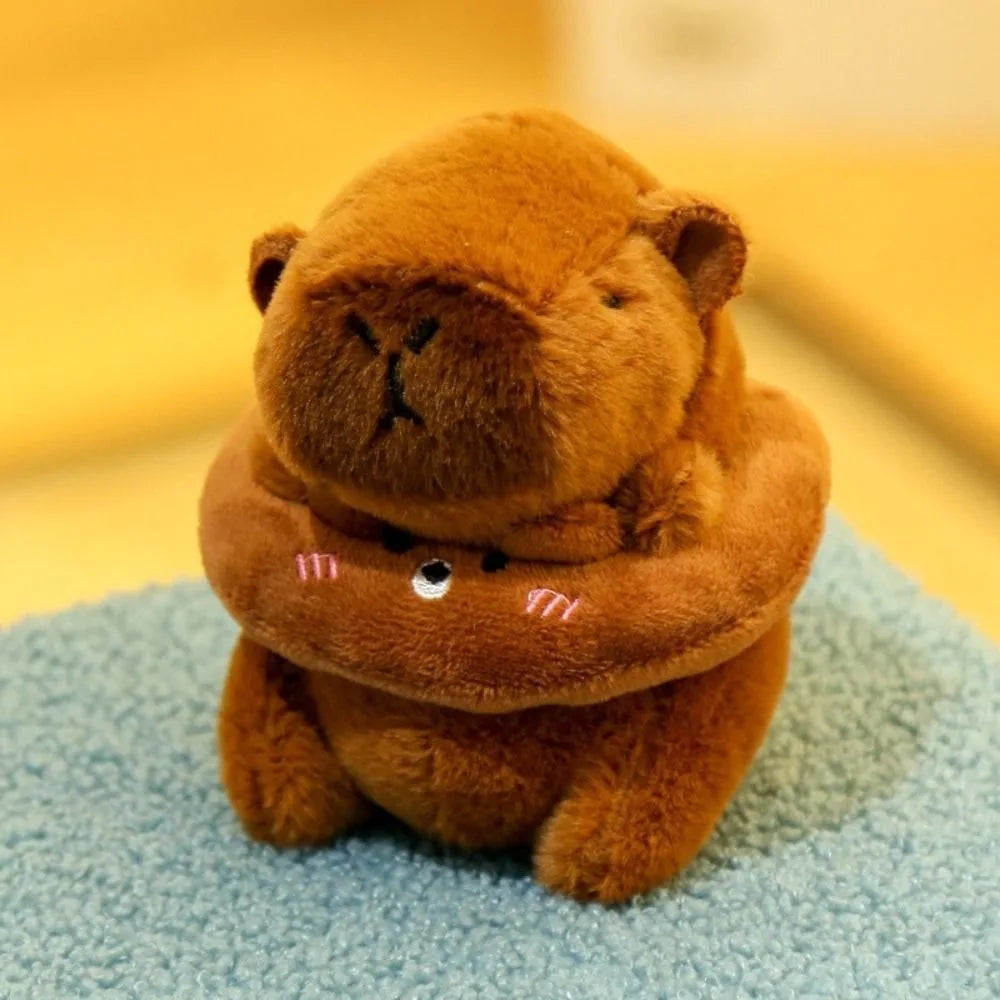 Flauschiger brauner Capybara-Plüschtier mit Hoodie und kleinen Flügeln, sitzt auf einer hellblauen Oberfläche.