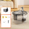 A clear pet drinking fountain with a metal spout is shown on a beige fabric surface.