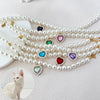 A strand of faux pearl necklaces featuring colorful heart pendants, displayed next to a white cat.