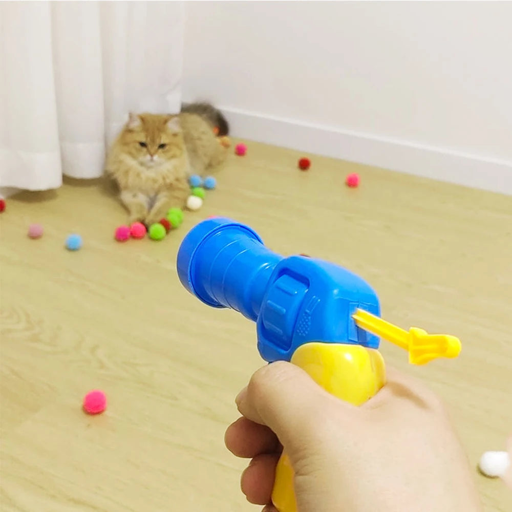 A hand is holding a blue interactive cat toy gun, aiming it at soft plush balls on a wooden floor.