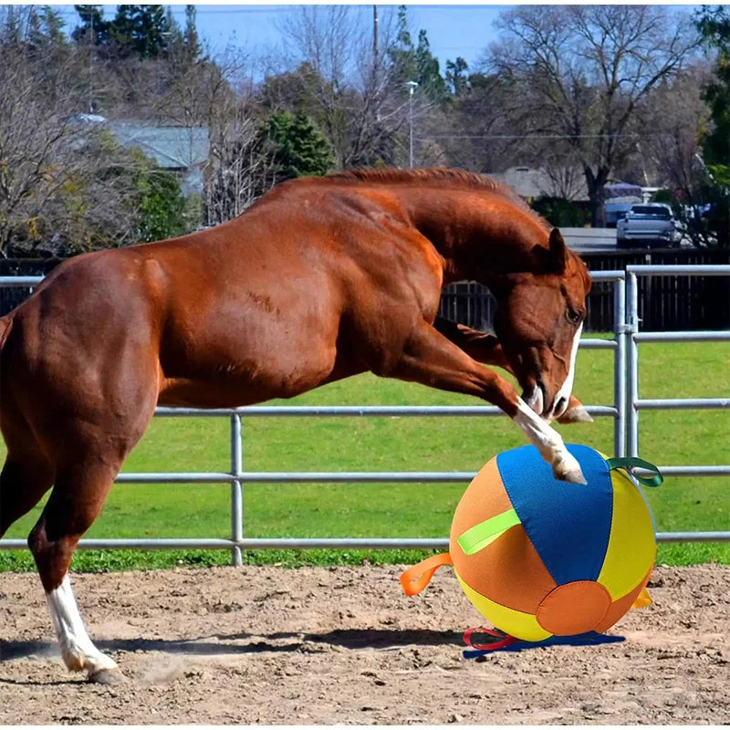 A brown horse is positioned next to a large, inflatable orange and blue herding ball on a dirt field.