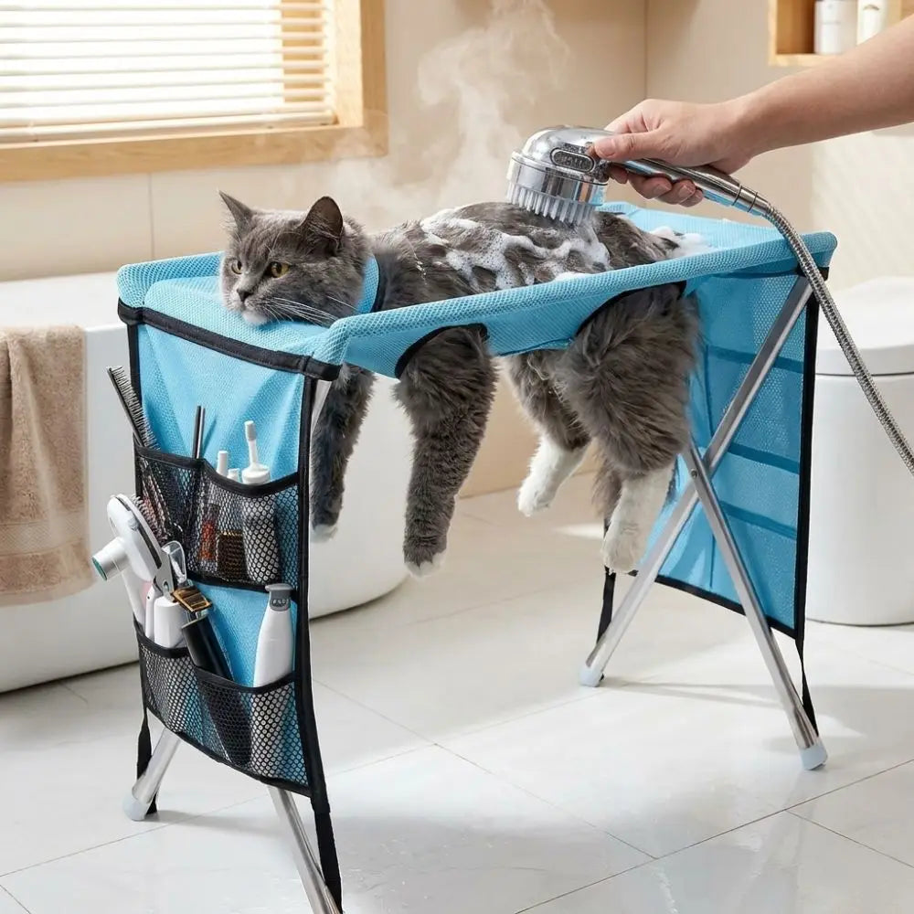 A cat is being groomed using a folding pet grooming hammock with a sturdy support frame and a handheld brush.