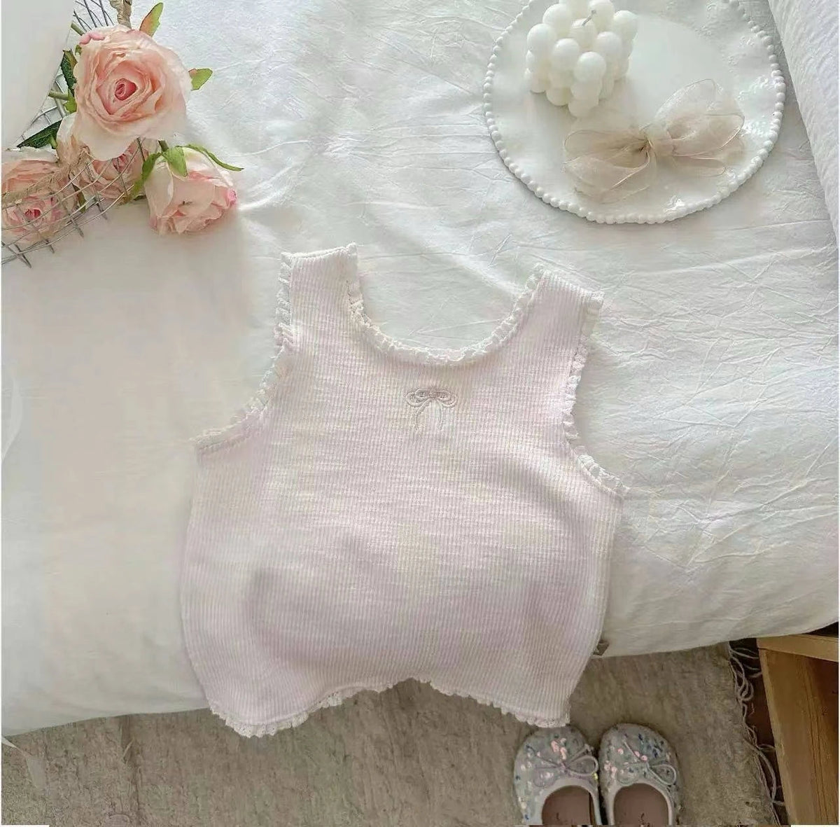 A light pink knitted mini vest with lace trim is displayed on a white surface next to roses and a bow.