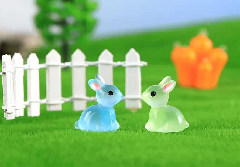 A set of two glow-in-the-dark resin bunny figurines, one blue and one green, placed on bright green grass behind a white pick