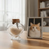 A clear glass jar with a heart shape filled with pet hair and topped with a feather, next to a framed photo of an orange tabb
