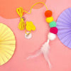 A colorful hanging cat toy featuring pom-poms and a feather on a yellow rope against a pink background.
