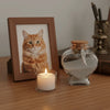 A glass jar filled with pet hair sits next to a lit candle and a framed cat photo on a wooden surface.