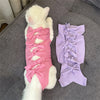 A white cat is lying next to a purple, lacy post-operative recovery vest for cats.