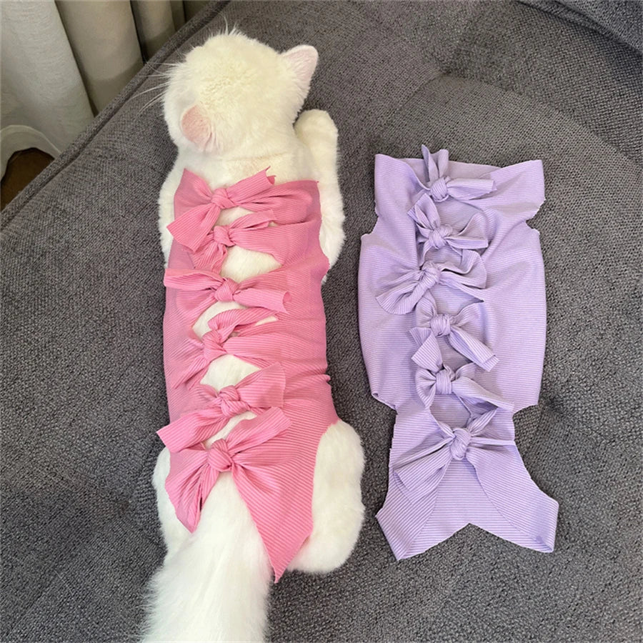 A white cat is lying next to a purple, lacy post-operative recovery vest for cats.