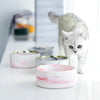 A white marbled ceramic double pet bowl set with a wooden stand sits on a white surface next to a cat.