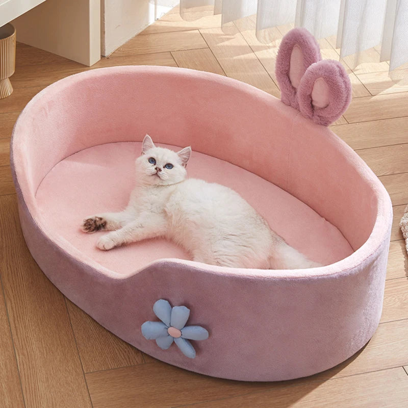 A pink plush pet bed for a kitten is shown on a wooden floor with a fluffy pink ear accessory.