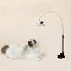 A white cat is lying down while an interactive feather wand toy with a bell extends above it.