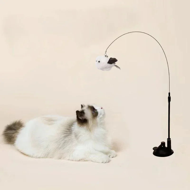 A white cat is lying down while an interactive feather wand toy with a bell extends above it.