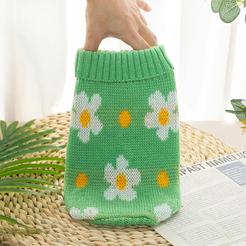 A hand is holding a bright green knitted dog sweater with white and yellow floral patterns on a woven mat.