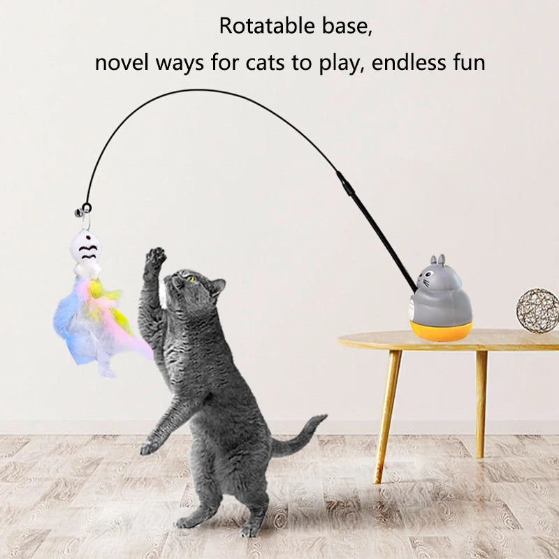 Eine graue Katze spielt mit einem rotierenden Stabspielzeug mit Feder und Figur auf einem Holztisch.