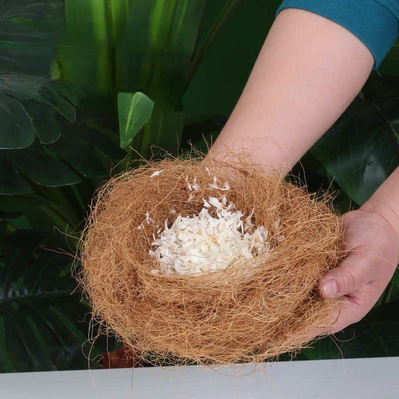 A hand holds a nest made of natural coconut fiber bird bedding against a dark green tropical leaf background.