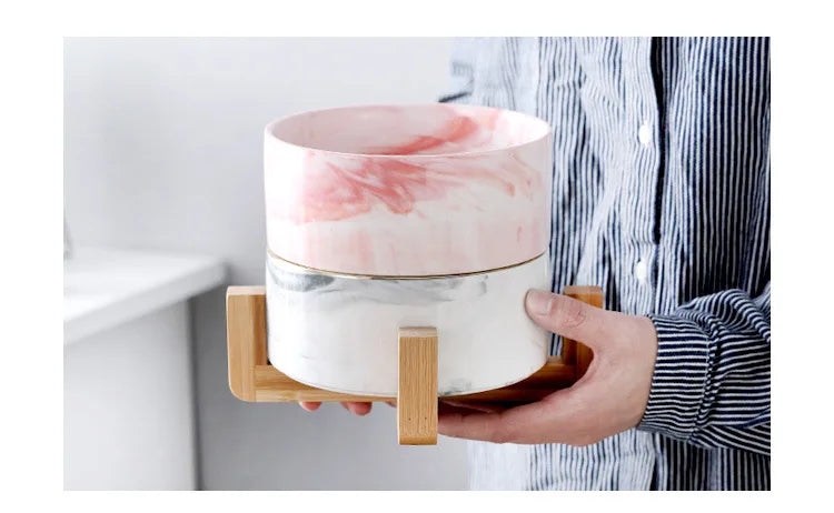 A pink and white marbled ceramic double pet bowl set with a wooden stand being held by a person.