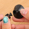 Una mano sostiene un anillo de alimentador para peces negro con múltiples formas y cebo de color verde azulado translúcido.