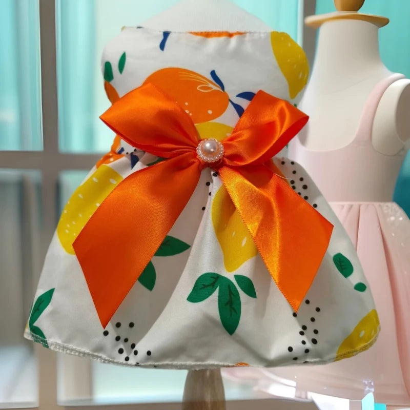A white dog dress with orange and yellow fruit print and an orange satin bow displayed on a mannequin.