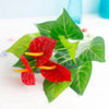 A vibrant red artificial anthurium fake plant with green leaves and yellow spathes on a white surface.