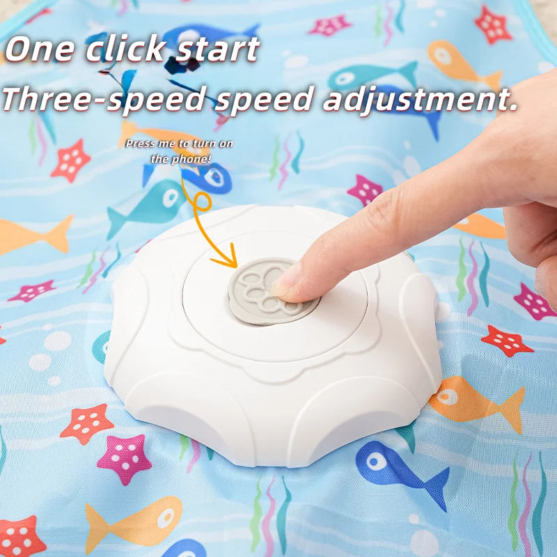Une main appuie sur un bouton d'un jouet interactif pour chat blanc posé sur un tissu bleu avec des poissons colorés.