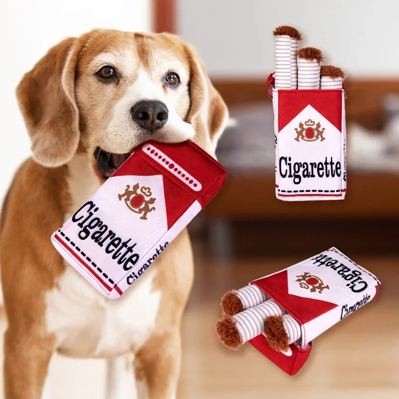 A dog holding a red and white cigarette box toy with cylindrical items inside.