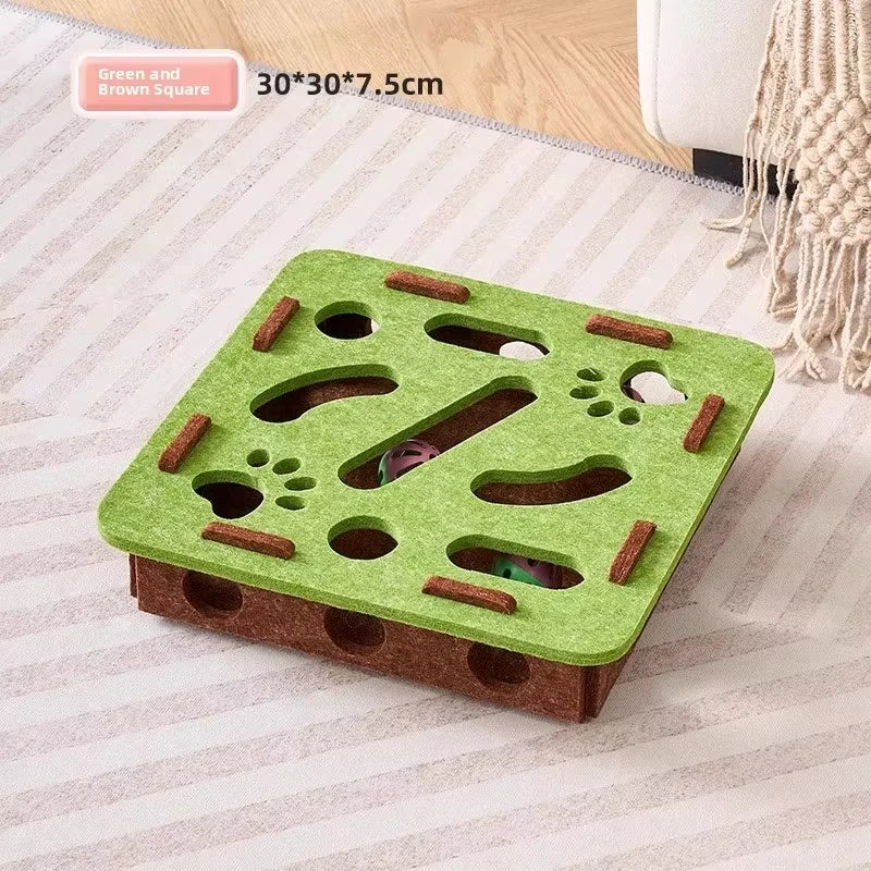 A green and brown square felt puzzle toy with holes and slots for cats is shown on a light fabric surface.