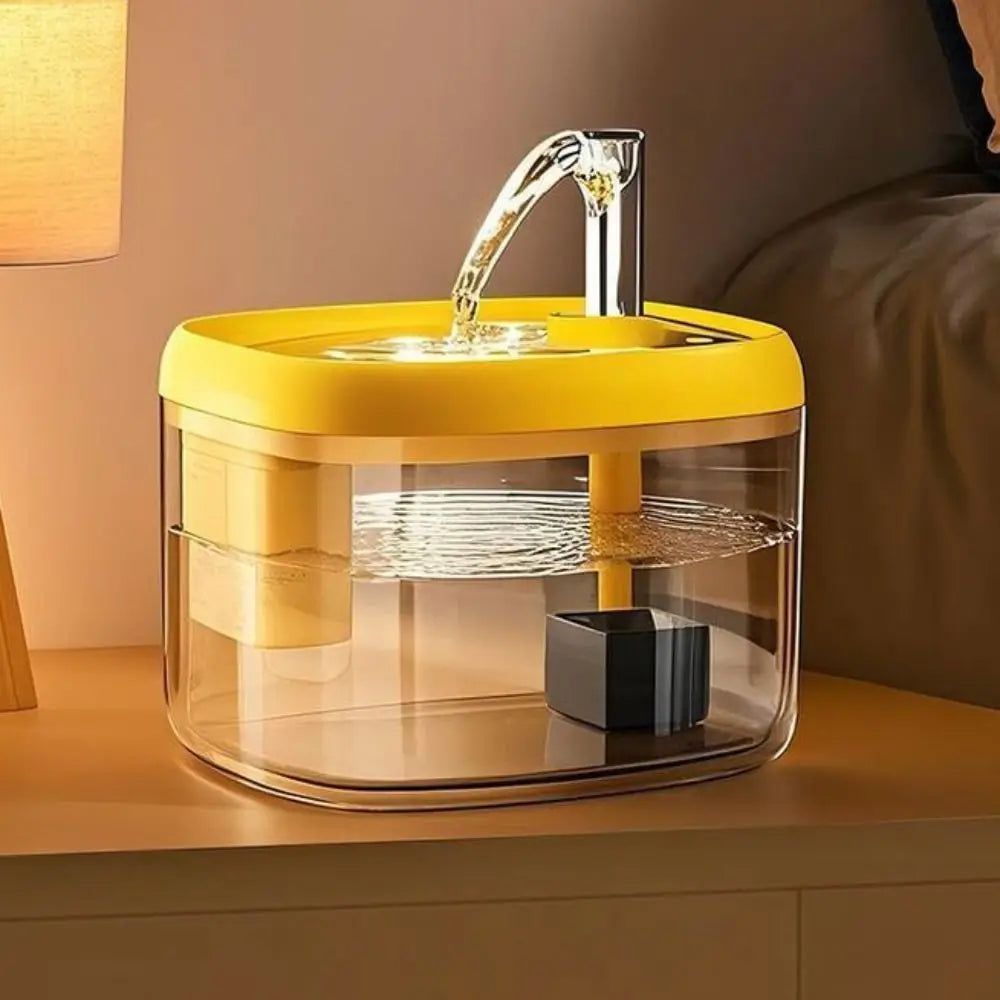 A yellow pet drinking fountain with a clear glass bowl and a metal spout is displayed on a wooden surface.