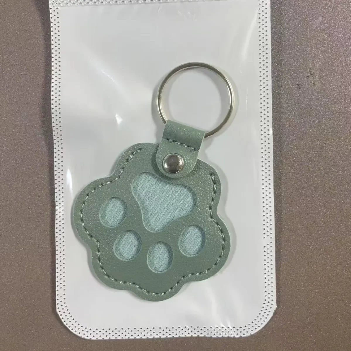 A light green leather pet fur keepsake keychain charm is displayed on a clear plastic pouch.