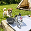 A clear glass water bottle with a carabiner clip rests on a white tray outdoors near a dog and a tent.
