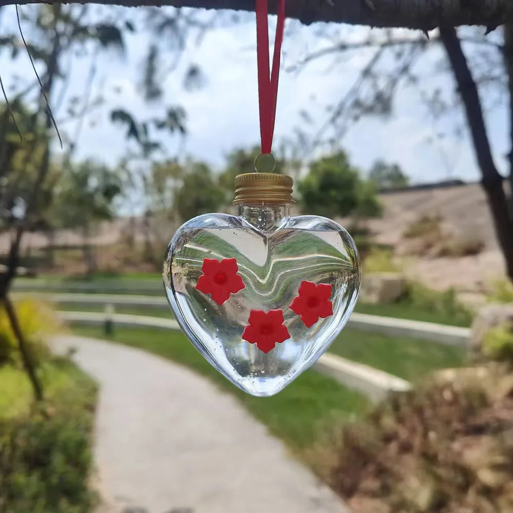 A clear glass heart-shaped hanging feeder with a red flower decoration suspended by a red string in an outdoor setting.