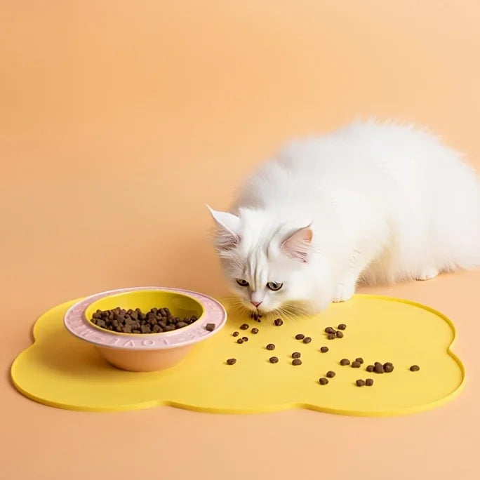 A white cat is eating dry pet food from a yellow silicone feeding mat.