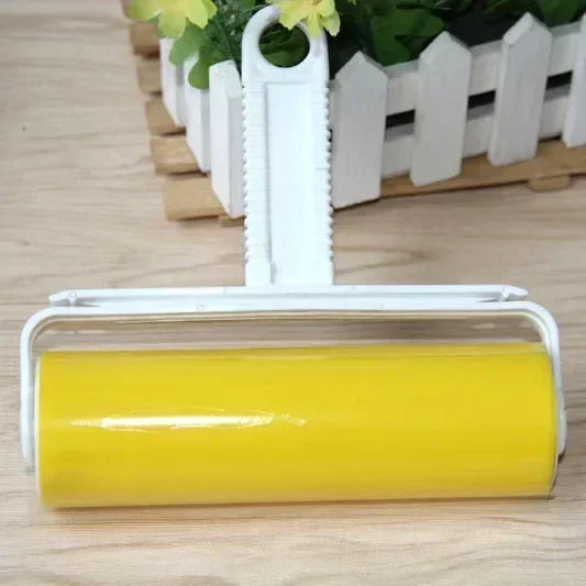 A white reusable lint roller with a yellow roller is shown in front of a wooden crate and greenery.