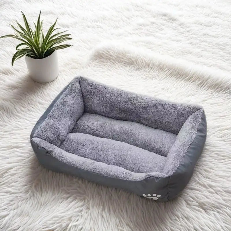 A gray plush dog bed is placed on a white fluffy surface next to a potted plant.