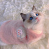 A fluffy white and blue cat wearing a pink embroidered vest on a white fur background.