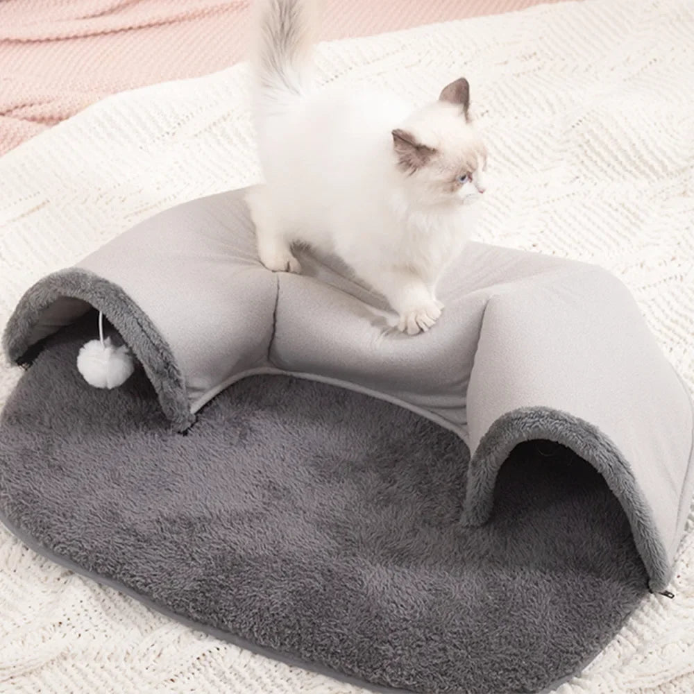 A white kitten sits inside a gray oval cat tunnel and cozy bed nest on a white textured blanket.