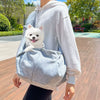 A person wearing a white hoodie is carrying a small white dog in a light blue hands-free pet sling carrier.