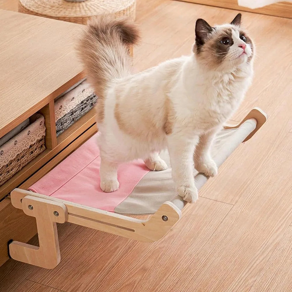 A fluffy calico cat is sitting on a light wood window perch designed for climbing and resting.