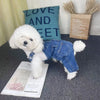 A white dog wearing a denim jumpsuit costume is positioned next to a sign and some dried grasses.