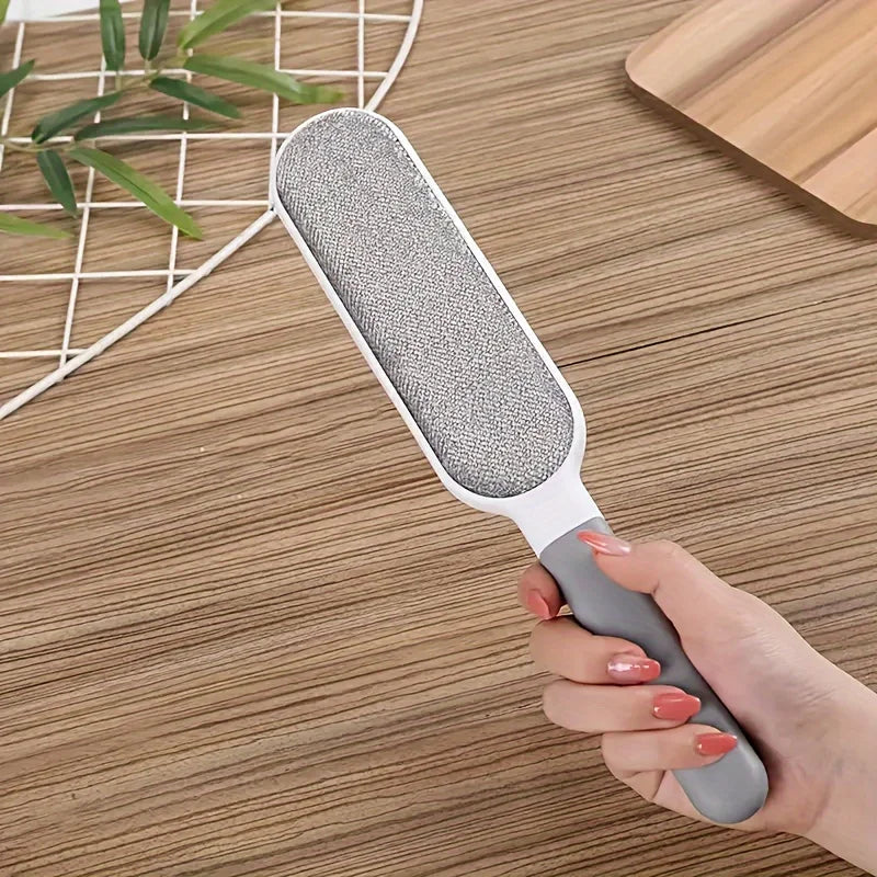 Une main tient une brosse retire-poils à double face avec une tête grise et un manche gris foncé sur une surface en bois.