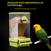 A yellow bird is feeding from a green automatic food replenishment feeder with a transparent window.
