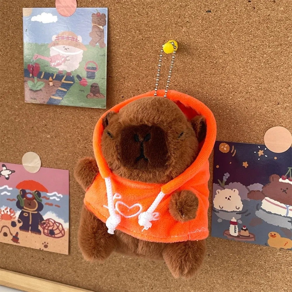 Orangefarbener Capybara-Plüsch-Hoodie mit kleinen Flügeln neben farbenfrohen Karten auf brauner Oberfläche