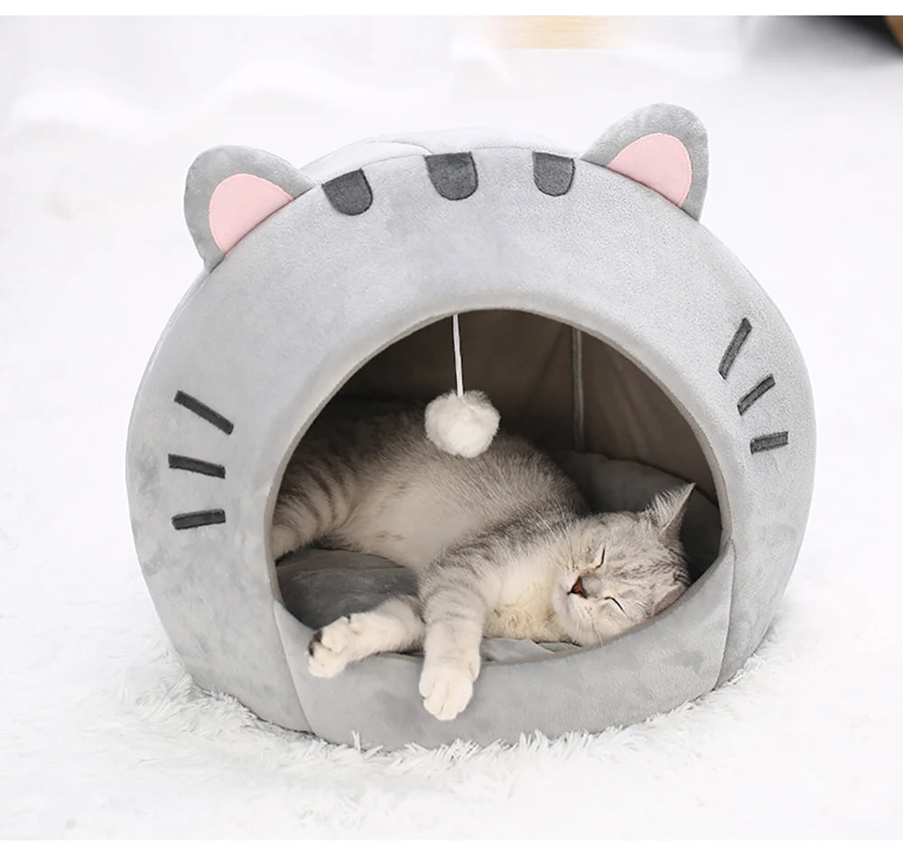 Un lit grotte en forme de chat gris avec des oreilles roses, contenant un chaton tigré endormi.