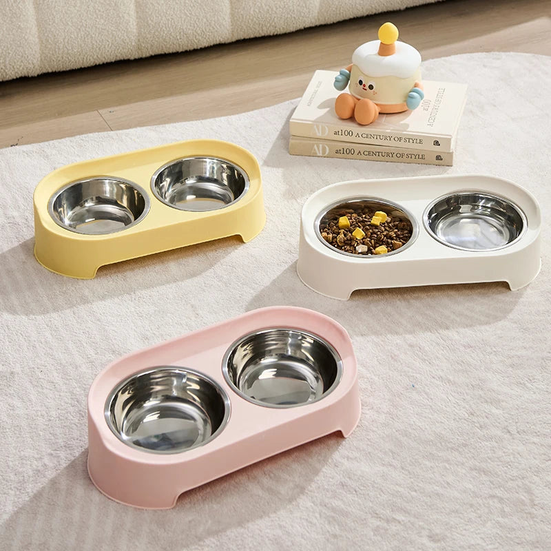 A set of three elevated pet feeders with double stainless steel bowls in yellow, white, and pink on a light fabric surface.