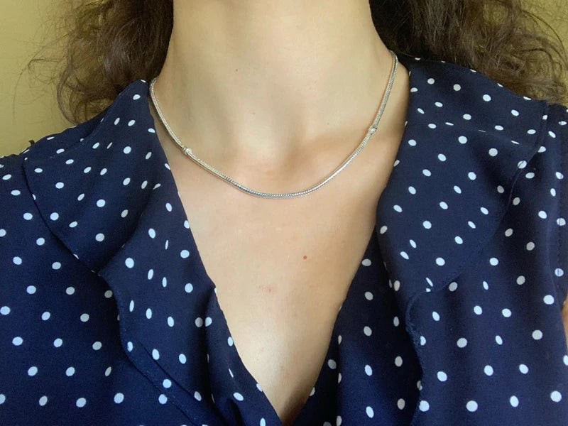 A close-up of a woman wearing a silver snake chain necklace with a heart charm over a dark blue polka dot top.