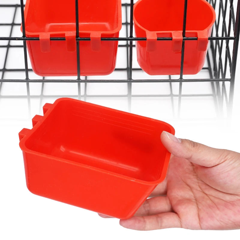 A hand holds a bright red plastic bird feeder and water bowl hanging in a black wire cage.