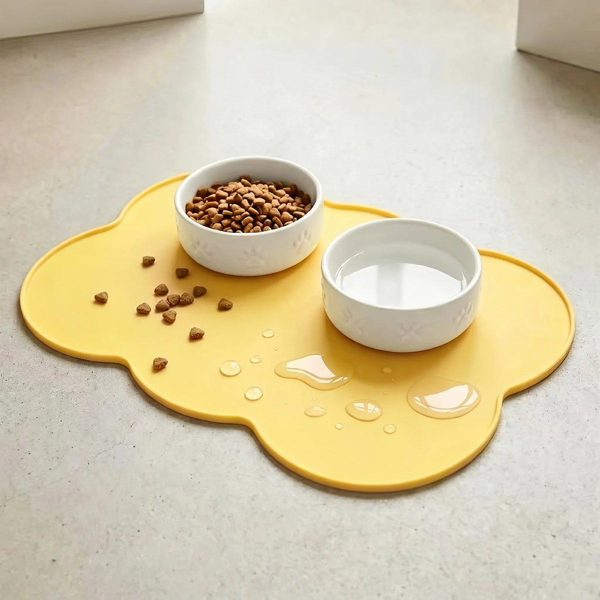 A yellow silicone pet feeding mat with raised edges holds a white bowl of dry kibble and a bowl of liquid food.